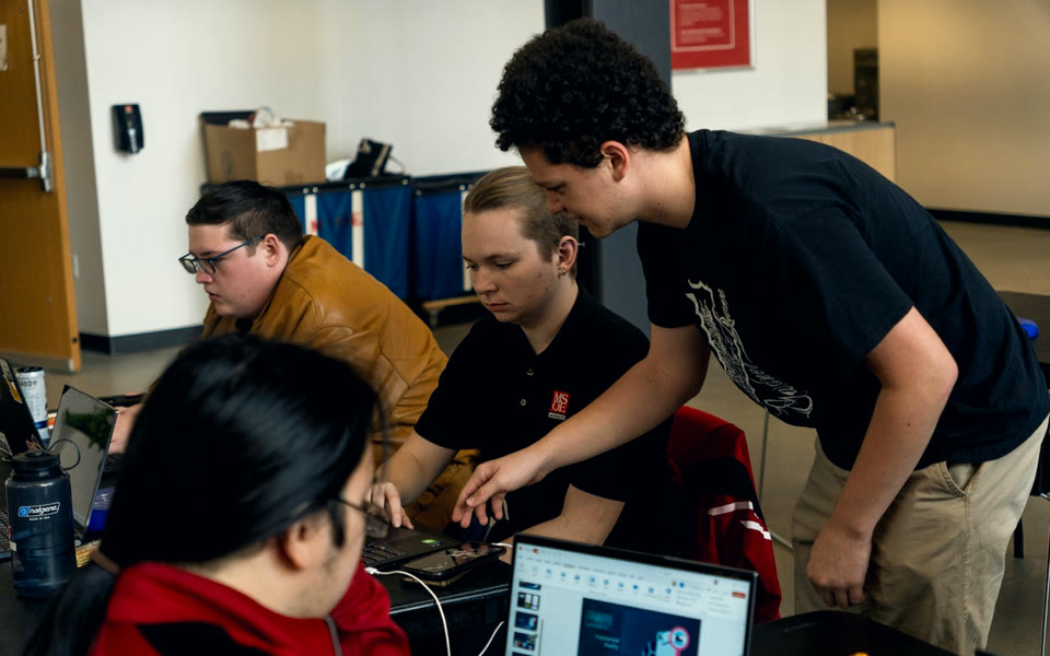 A student leans in to help a team working at their laptops during a 2024 hackathon.