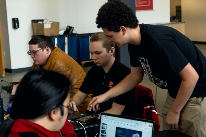 A student leans in to help a team working at their laptops during a 2024 hackathon.